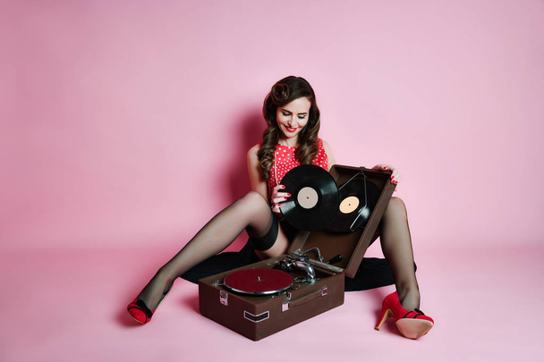 Glamorous pinup girl sitting near retro gramophone holding LP vinyl record looking up on pink background - 写真・画像