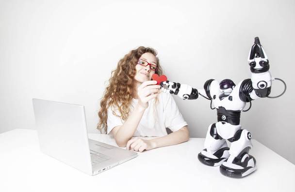 lächelnde rothaarige Frau arbeitet mit Laptop vor weißer Wand, während Roboter ihr rotes Herz schenkt - Foto, Bild