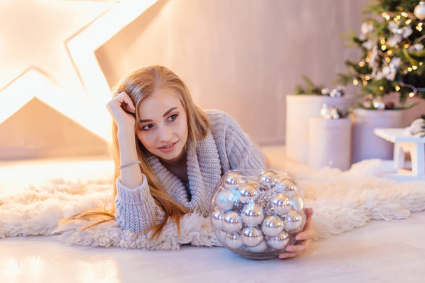 Beautiful young blond woman dressed in white sweater laying next to the big star with bowl full of silver balls in a holiday interior with Christmas tree. - Foto, Bild