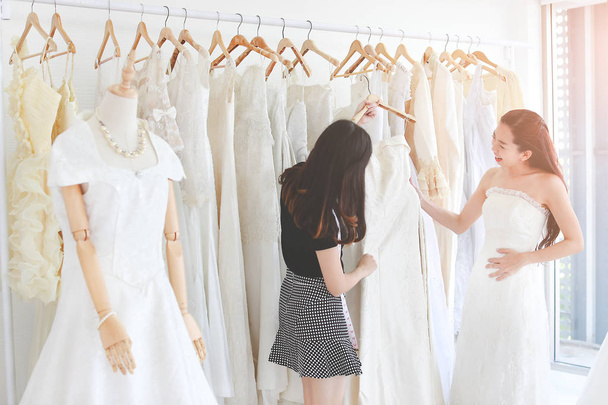 美しいアジアの女性のフィッティングとウェディング ドレスのデザイナーを支援を比較します。白のドレスの背景とコピー スペース. - 写真・画像