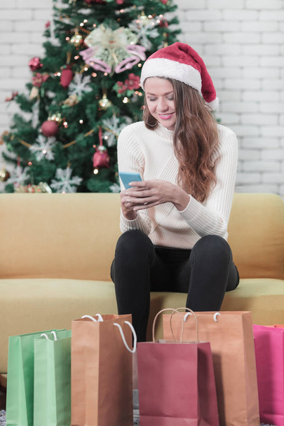 junge schöne kaukasische Frau mit rotem Hut, die auf einem Sofa vor Weihnachtsbäumen sitzt und auf das Smartphone und mehrere große Einkaufstüten vor sich blickt. Konzept für Ausgaben beim Weihnachtsfest. - Foto, Bild