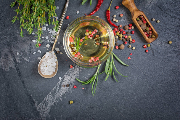 Aromatic or flavored olive oil in glass bowl with spices and herbs as chili peppers, rosemary and thyme on black marble background with copy space - Photo, Image