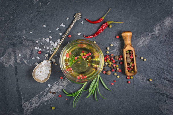 Aromatic or flavored olive oil in glass bowl with spices and herbs as chili peppers, rosemary and sea salt in silver spoon on black marble background - Fotografie, Obrázek