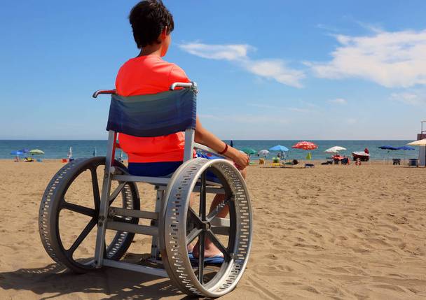 ελπιδοφόρα ΑμεΑ αγόρι σε μια αναπηρική καρέκλα κοιτάζει το γαλάζιο της θάλασσας - Φωτογραφία, εικόνα