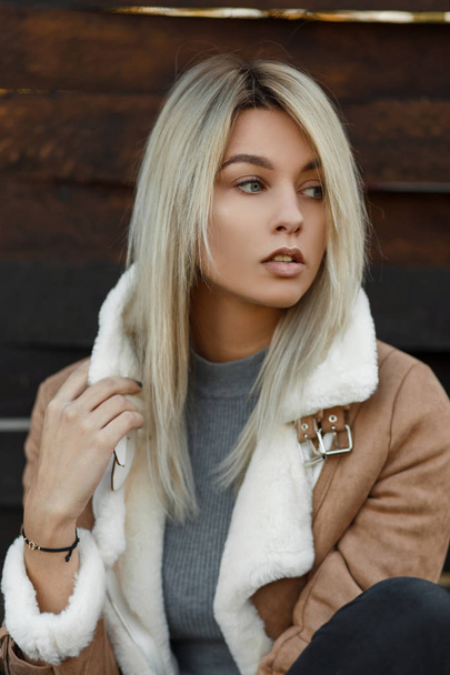 Serious pretty young woman with a natural make-up in a stylish autumn-spring jacket with a white fur against the background of a wooden wall, Portrait of a beautiful girl. - 写真・画像