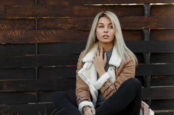 Mulher bonita nova em uma jaqueta bege de estação de moda com um colarinho de pele e suéter cinza senta-se na grama perto de uma cerca de madeira. Look moderno da moda. Menina atraente
 - Foto, Imagem