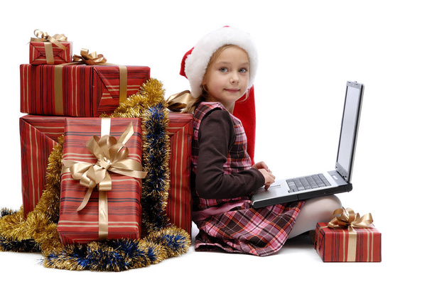 Little girl in Santa Claus cap with a laptop and Christmas gifts on white background, isolated - Foto, Bild