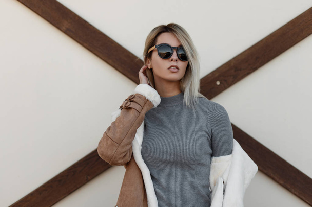 Elegante retrato de una joven chica bastante hipster en gafas de sol de moda y suéter vintage gris de moda con chaqueta de otoño de pie cerca de la pared blanca con madera
 - Foto, Imagen
