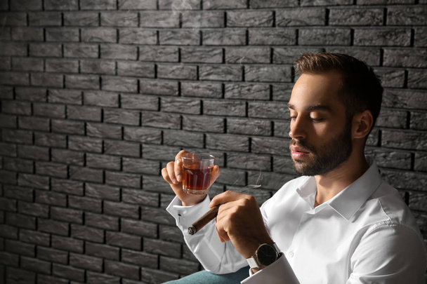 Jonge zakenman met glas whiskey en sigaar in de buurt van donkere bakstenen muur - Foto, afbeelding