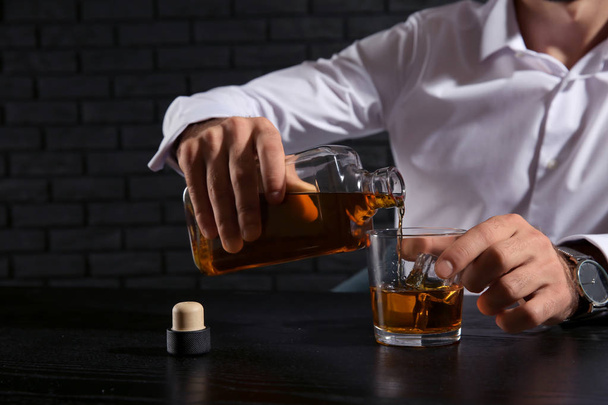 Mann gießt Whisky ins Glas, während er am Tisch auf dunklem Hintergrund sitzt - Foto, Bild