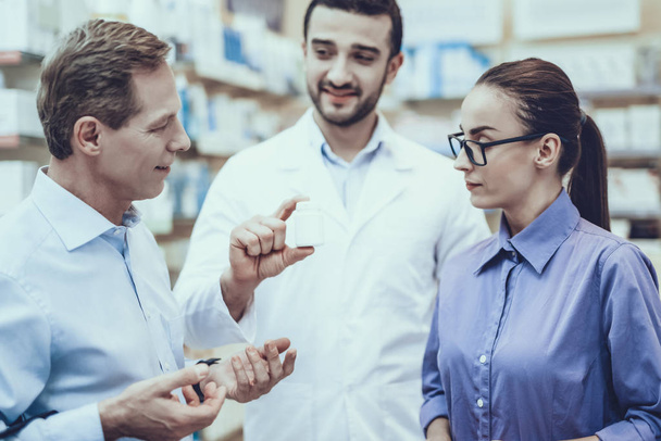 Customers Choose a Medicaments. Pharmacist Showing a Bottle with Pills to Customers. Customers is Adult Man and Young Woman. Pharmacist Wearing a Special Medical Uniform. People Located in Pharmacy. - Φωτογραφία, εικόνα