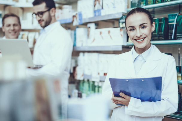Pharmacists is Working. Woman Writing Information about a Medicine. Men on Background Using a Laptop. People Wearing a Special Medical Uniform. Woman is Smiling. People Located in Pharmacy. - Foto, afbeelding