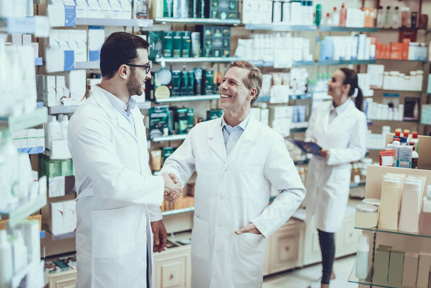 Pharmacists is Working. Two Men Handshaking with Each Other. Persons is Smiling. Woman Writing Information about a Medicine. People Wearing a Special Medical Uniform. People Located in Pharmacy. - Фото, изображение