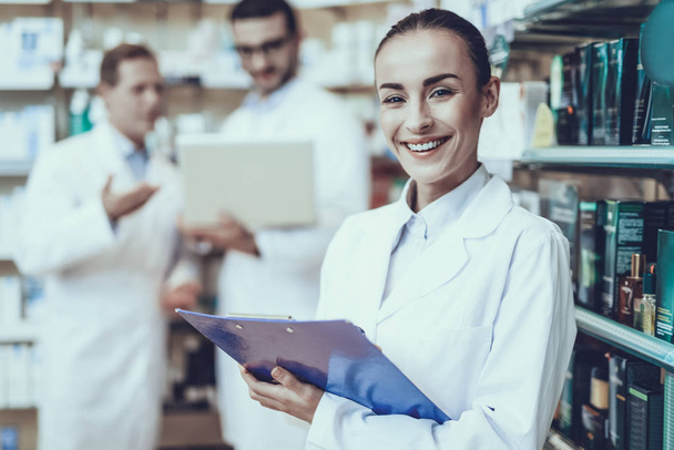 Pharmacists is Working. Woman Writing Information about a Medicine. Men on Background Using a Laptop. People Wearing a Special Medical Uniform. Woman is Smiling. People Located in Pharmacy. - Photo, Image