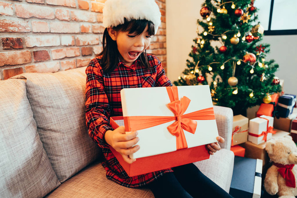 little cute girl open christmas gift box with a surprised face. cheerful kid excited looking at xmas present. decorated room with tree and teddy bear for coming up holidays winter vacation new year. - Foto, Bild