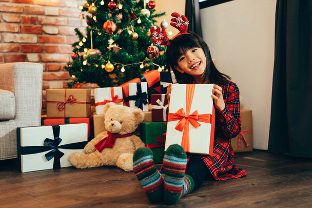 cute daughter face camera showing big gift box enjoy smiling sitting on ground near decorated Christmas tree and teddy bear. attractive girl childhood with reindeer got xmas present from santa claus. - 写真・画像