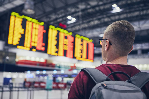 Achteraanzicht van de jongeman die de vluchtschema 's controleert op het aankomstvertrekbord op de luchthaven. - Foto, afbeelding