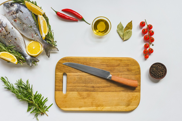 flat lay with wooden board, knife, fish and ingredients on white table - Foto, Imagem