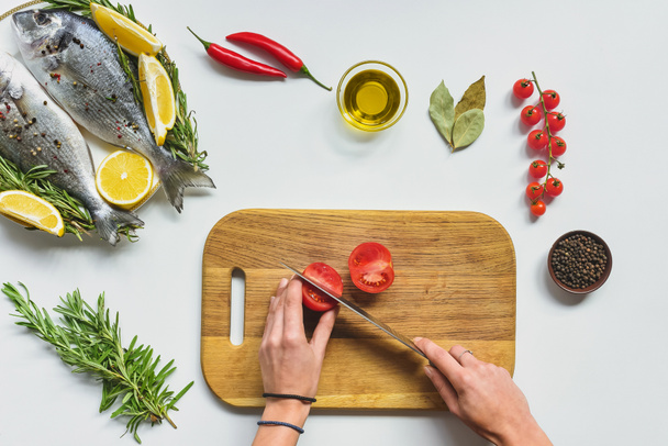 imagen recortada de mujer cortando tomate por cuchillo en tablero de madera cerca de pescado crudo en la mesa
 - Foto, imagen