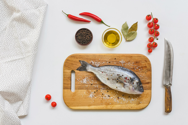 flat lay with uncooked fish on wooden board and various ingredients on white table - Zdjęcie, obraz