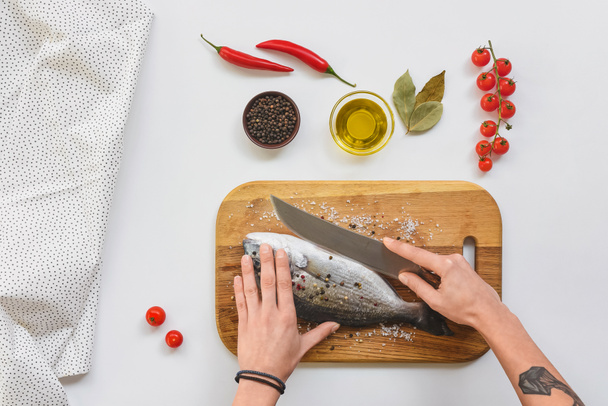 immagine ritagliata di donna tatuata che taglia il pesce con un coltello su tavola di legno vicino agli ingredienti sul tavolo
 - Foto, immagini