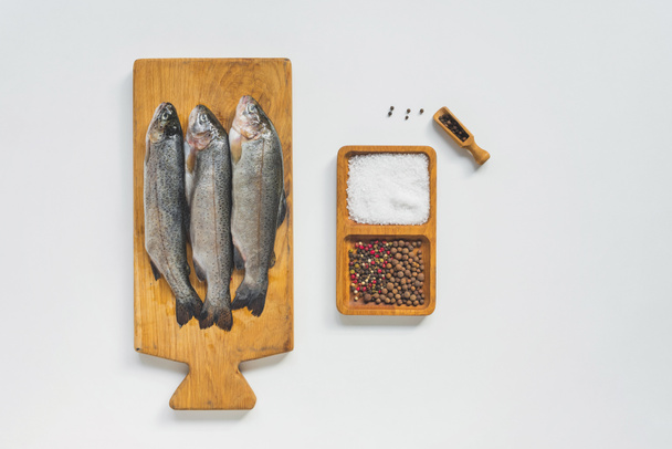 view from above of uncooked fish on wooden board near salt and pepper on white table  - Valokuva, kuva