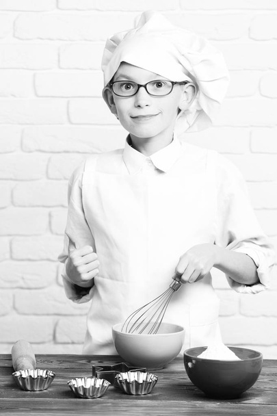 young boy small cute cook chef in white uniform and hat on smiling face with glasses standing near table with rolling pin and colorful bowls and cooking on brick wall background - Photo, Image