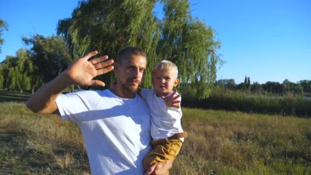 幸せな父と幼い息子が公園でカメラに手を振っての肖像画。若いお父さん夏自然に腕の中で彼の子を保持します。一緒に時間を過ごす家族屋外。スローモーションをクローズ アップ. - 映像、動画