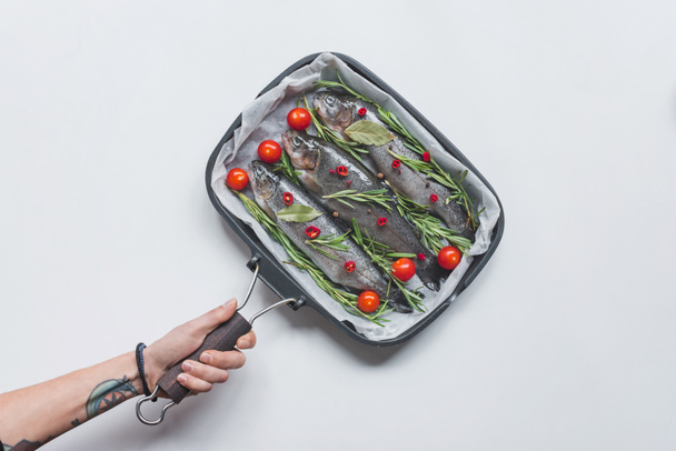 imagen recortada mujer sosteniendo bandeja con pescado con romero, hojas de laurel y tomates cherry sobre mesa blanca
 - Foto, imagen