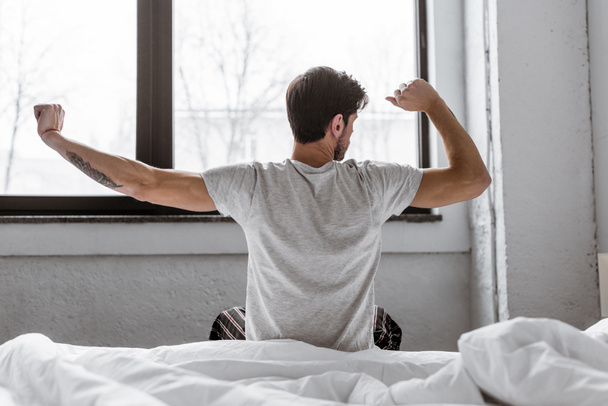visão traseira do jovem de pijama que se estende enquanto está sentado na cama pela manhã
 - Foto, Imagem