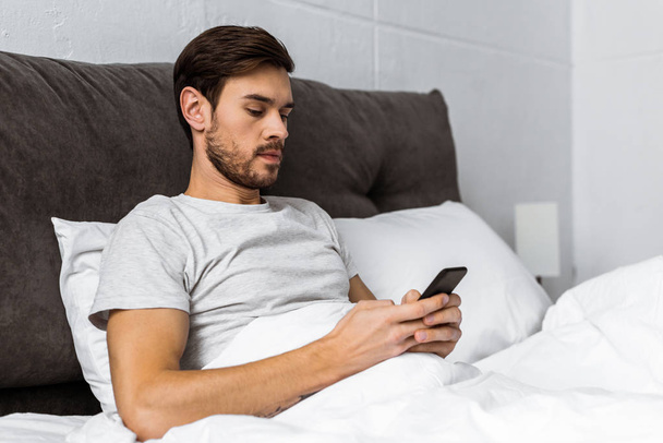 handsome bearded young man using smartphone in bed in the morning - Foto, Imagem