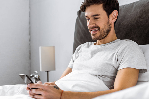 handsome smiling young man sitting in bed and using smartphone - Photo, Image
