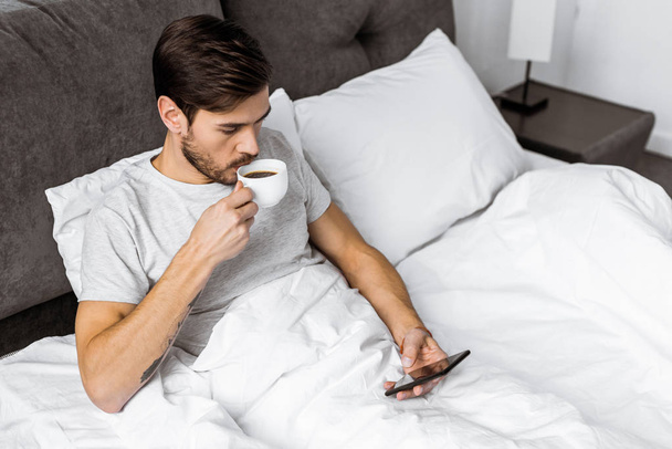 high angle view of young man using smartphone and drinking coffee in bed - Foto, imagen