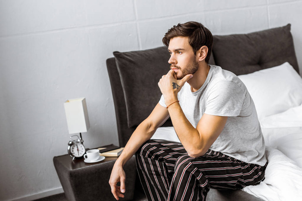 thoughtful young man in pajamas sitting on bed and looking away in the morning - Foto, immagini