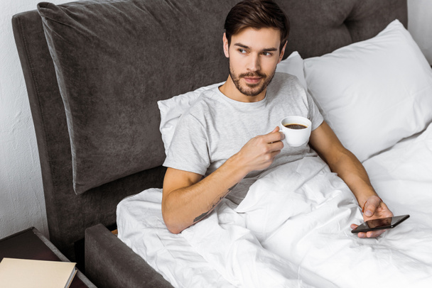 若い男がベッドでスマート フォンを使用し、一杯のコーヒーを保持のハイアングル - 写真・画像