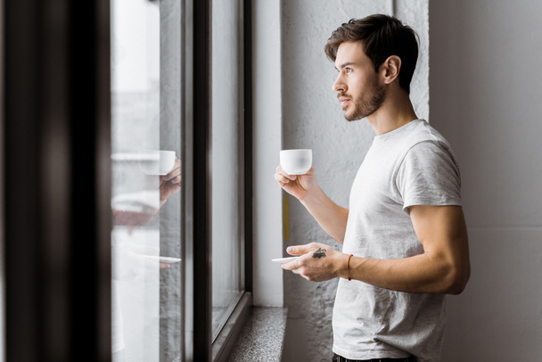 вид збоку молодого чоловіка тримає чашку кави і дивиться на вікно вранці
 - Фото, зображення