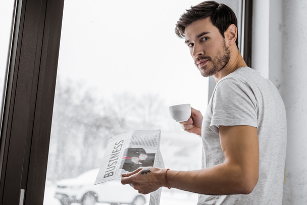 joven guapo con taza de café y periódico mirando a la cámara mientras está de pie cerca de la ventana
 - Foto, Imagen