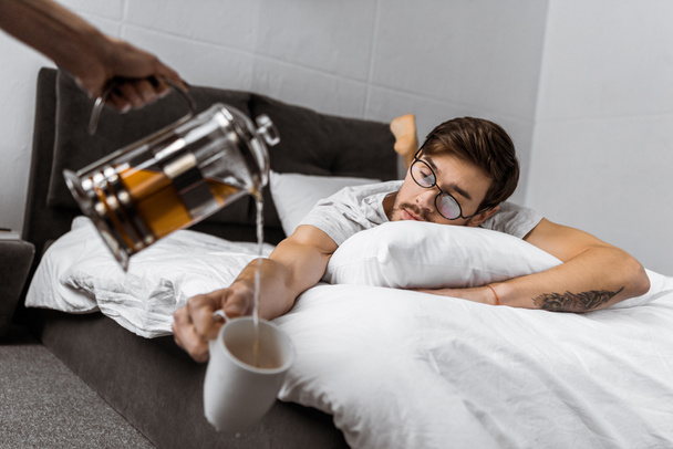 partial view of someone pouring tea into cup while sleepy man in eyeglasses lying on bed   - Foto, Imagem