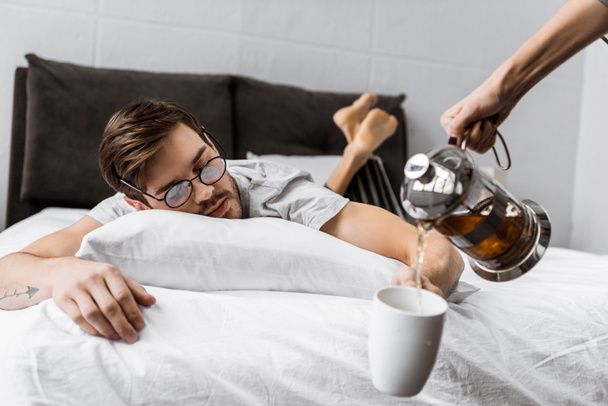 plan recadré de quelqu'un versant du thé dans une tasse tandis que l'homme endormi dans des lunettes couché sur le lit
  - Photo, image