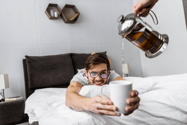 cropped shot of someone pouring tea to smiling man in eyeglasses lying on bed and holding cup - Photo, Image