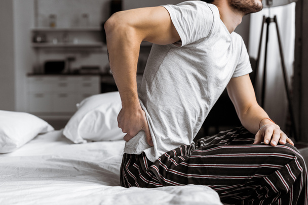 обрезанный снимок молодого человека в пижаме, страдающего от боли в спине, сидя на кровати утром
 - Фото, изображение