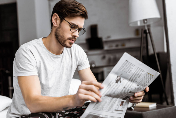 young man in pajamas and eyeglasses sitting on bed and reading newspaper in the morning - Foto, immagini