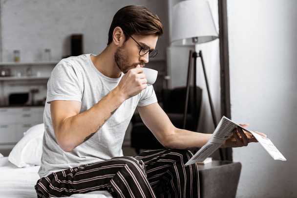 Nuori mies pyjamassa ja silmälasit juomassa kahvia ja lukemassa sanomalehteä aamulla
 - Valokuva, kuva