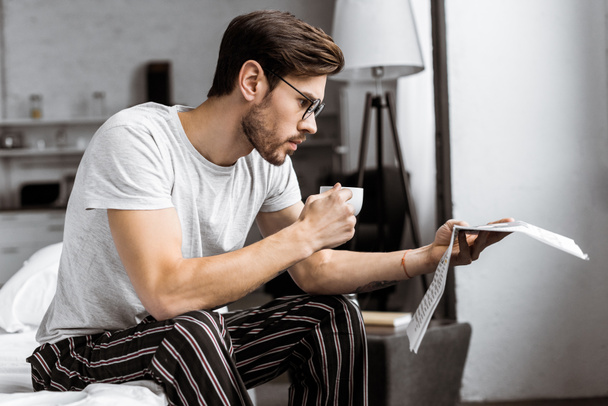 handsome young man in pajamas and eyeglasses holding cup of coffee and reading newspaper in the morning - Foto, imagen