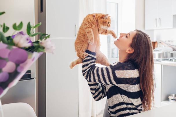 Nuori nainen leikkii kissan kanssa keittiössä kotona. Tyttö pitelee punaista kissaa. Onnellinen isäntä hauskaa lemmikkinsä kanssa
 - Valokuva, kuva