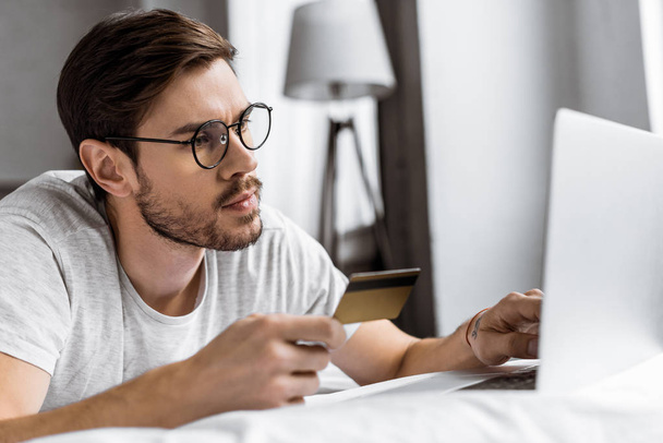 bel giovane uomo con gli occhiali in possesso di carta di credito e utilizzando il computer portatile sul letto
 - Foto, immagini