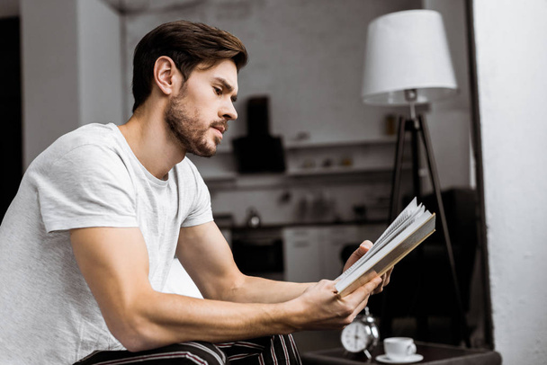 side view of handsome young man in pajamas sitting and reading book at home - Foto, imagen