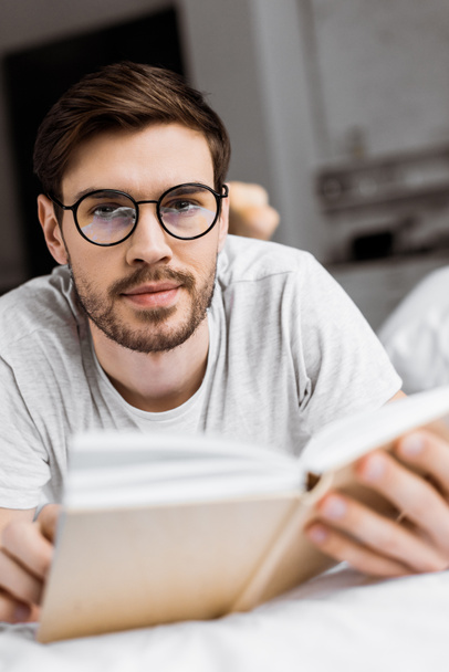 young man in eyeglasses holding book and looking at camera while lying on bed - Photo, Image