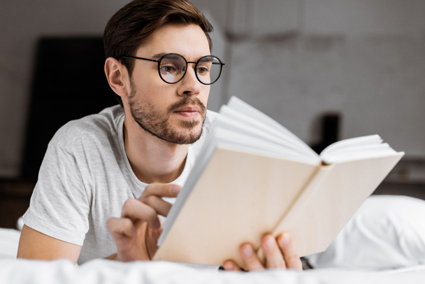 handsome young man in eyeglasses lying in bed and reading book  - Foto, Imagem