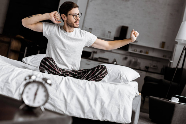 pohledný mladý muž v pyžamu a brýle, strečink, zatímco sedí na posteli v noci - Fotografie, Obrázek